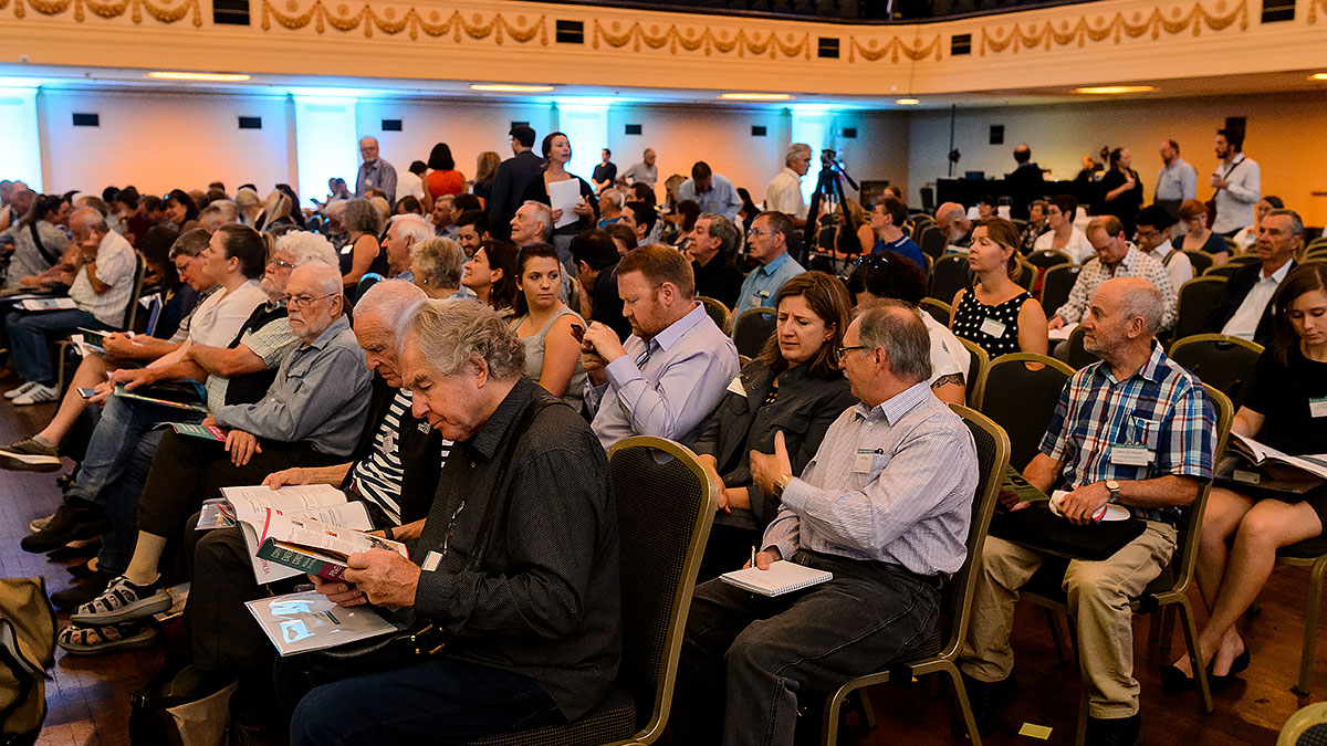 A large hall full of people sitting on chairs at an event, talking with each other, reading their programs or looking towards the front of the hall.
